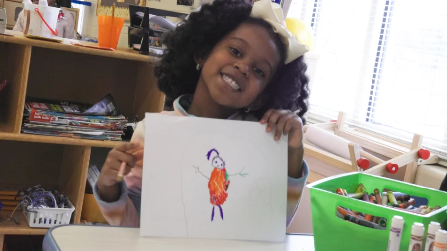 A young girl with curly black hair, wearing a large cream-colored bow, smiles brightly while holding up her drawing. The drawing features a stick figure with an orange body and purple hat. She is seated at a table in a classroom, with a green plastic bin filled with markers and crayons beside her. Shelves in the background contain books, art supplies, and a small basket of yarn.