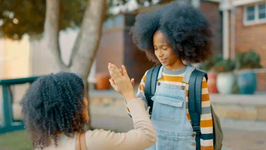 A young girl wearing a striped sweater, denim overalls, and a backpack smiles while giving a high-five to an adult woman with curly hair. The interaction takes place outdoors in a sunny setting, with trees, potted plants, and a brick building visible in the background.