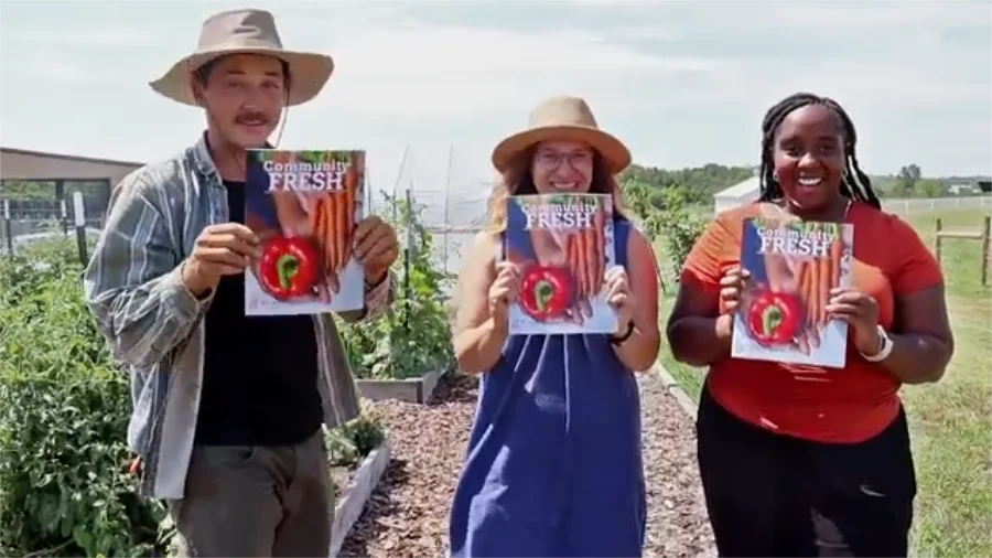 Three people stand in a community garden, each holding up a magazine titled "Community FRESH" featuring an image of carrots and a red bell pepper. The person on the left, a man with a mustache, wears a wide-brimmed hat, a black shirt, and an open denim jacket. The middle person, a woman in a straw hat and blue dress, smiles while holding the magazine. The person on the right, a woman wearing a bright orange shirt, grins while holding her copy. The background shows raised garden beds with green plants and a clear sky.