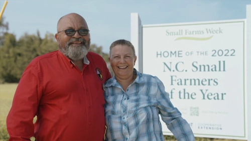 A man and woman standing outdoors, smiling, with their arms around each other. The man wears a red shirt and sunglasses, while the woman wears a blue plaid shirt. Behind them is a sign that reads: "Small Farms Week - Home of the 2022 N.C. Small Farmers of the Year," with additional text for "Cooperative Extension."