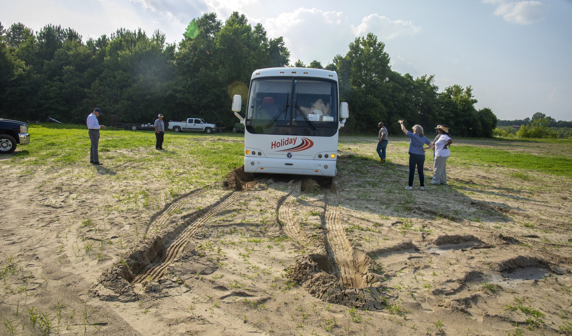 A white Holiday tour bus stuck in sandy ground, with deep tire ruts in front of it. Several people stand around the scene on the open field, some observing and one gesturing toward the bus. Pickup trucks are parked in the background near a treeline under a partly cloudy sky.