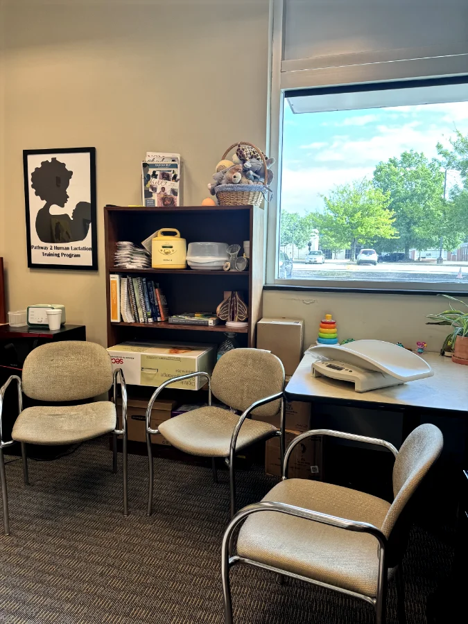 A lactation consultation office with three beige fabric chairs with metal frames arranged in a semi-circle. A wooden bookshelf holds a breast pump, baby bottles, educational books, and a basket filled with baby dolls and plush toys. A black and white poster on the wall displays the words "Pathway 2 Human Lactation Training Program" with an illustration of a mother and baby. A desk near a large window has a baby scale, a small stack of rainbow stacking rings, and a potted plant. Natural light streams through the window, showing parked cars and green trees outside.