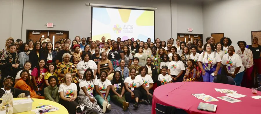 A large and diverse group of predominantly women, many wearing white t-shirts with a colorful logo, pose together in a conference room. They are smiling and looking at the camera. A projector screen in the background displays a colorful graphic with the words 'Uplifting Black Bodies Conference.' The room has bright lighting, with tables covered in red tablecloths in the foreground.