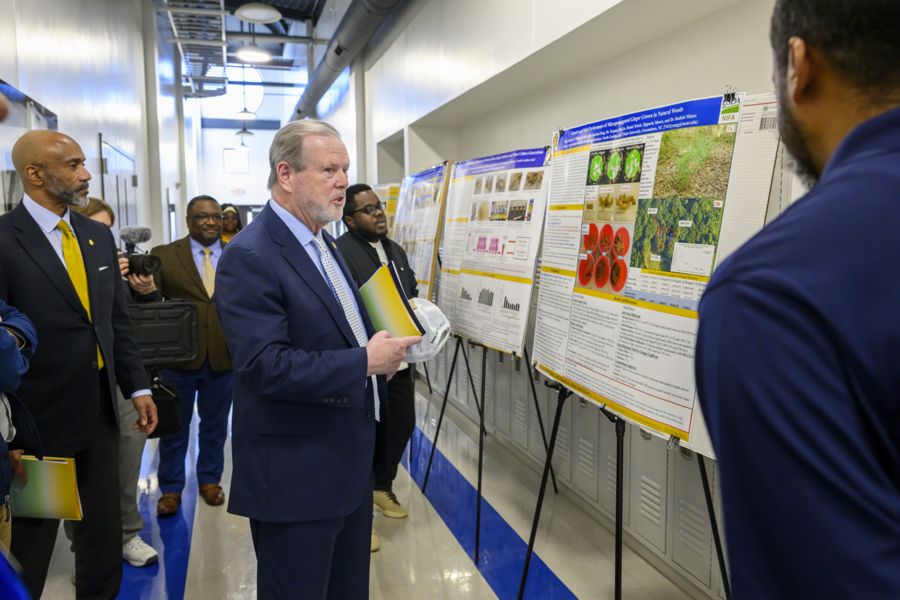 A group of professionally dressed individuals walk through a well-lit hallway, observing research posters displayed on easels. A man in a navy blue suit, holding a folder and a white construction helmet, closely examines one of the posters while speaking to another man in a dark blue shirt. Several other individuals in suits stand nearby, including a person holding a video camera. The posters feature images, graphs, and text, indicating a formal academic or research presentation setting.