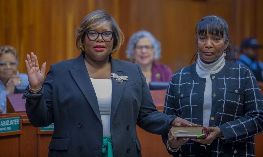 A woman wearing glasses and a dark green blazer raises her right hand while taking an oath of office. She has a confident expression and is standing in a formal setting with wooden paneling. Another woman, dressed in a black and white checkered jacket over a white turtleneck, holds a book with a serious expression. Behind them, individuals are seated, including one wearing a purple blazer, observing the ceremony.