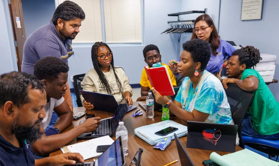 A diverse group of adults sits around a conference table in a classroom, engaging in a collaborative discussion. Several people are using laptops, one woman holds up a red notebook while speaking, and others listen attentively. The atmosphere appears focused and interactive, suggesting a group learning or training session.