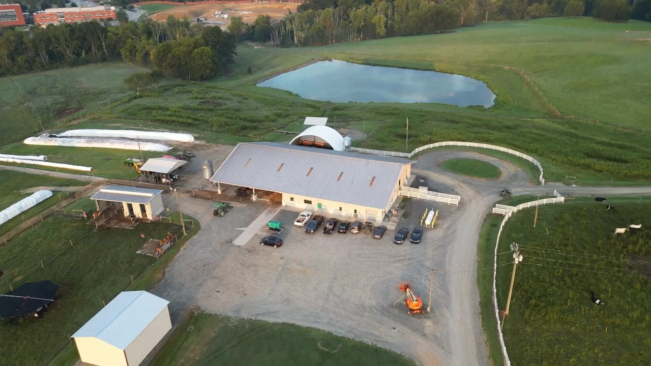 Aerial view of a modern dairy farm with multiple buildings, vehicles, and equipment. The central structure has a large metal roof with cars and farm machinery parked in front. Surrounding the main building are smaller sheds, fenced areas with livestock, and white fencing outlining the property. In the background, there is a large pond bordered by green pastures and wooded areas, with additional farmland visible in the distance.