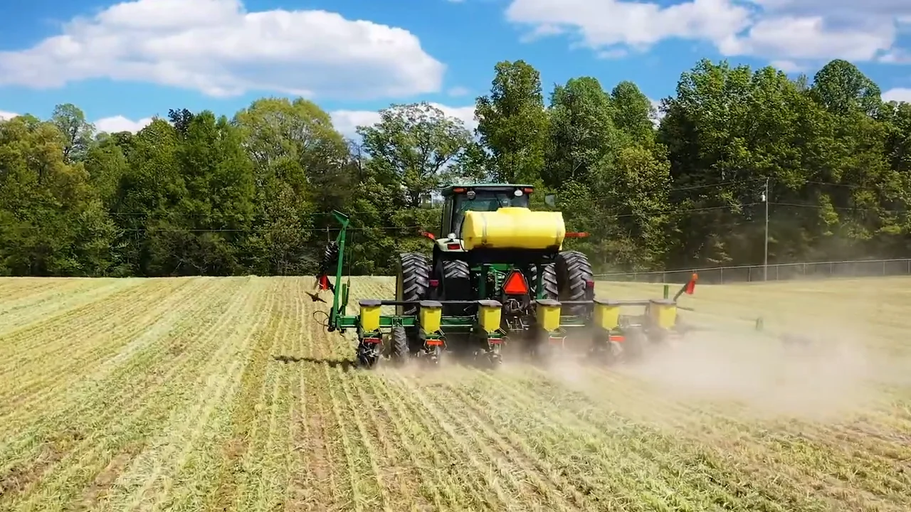 Rear view of a large green tractor with a yellow tank and attached planting equipment working in a freshly tilled field. The tractor is kicking up dust as it moves, creating a visible trail behind it. The field has evenly spaced rows, and the background features a dense line of green trees under a bright blue sky with scattered white clouds.